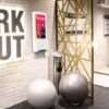 Digital hand sanitiser station mounted on a gym wall beneath a "WORK OUT" sign, positioned near the entrance to a fitness studio with equipment visible beyond.