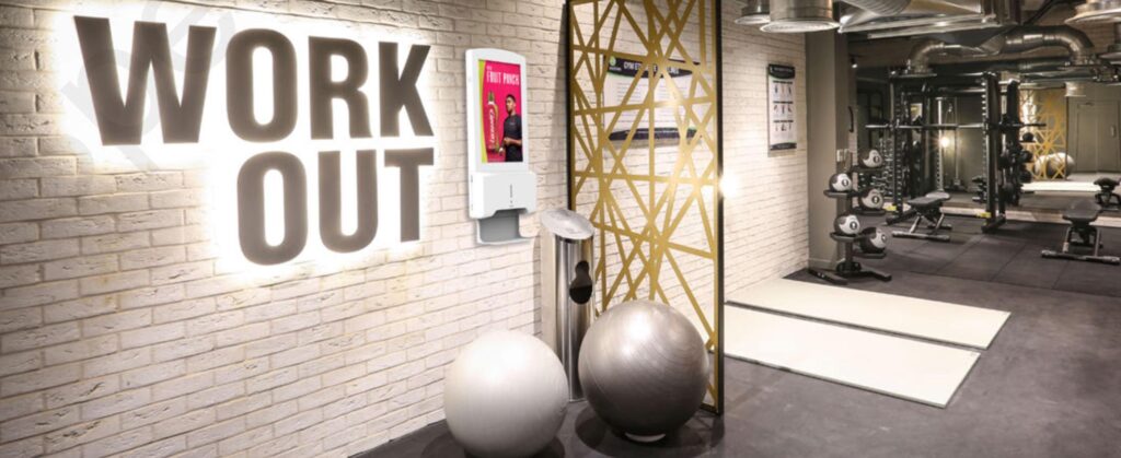 Digital hand sanitiser station mounted on a gym wall beneath a "WORK OUT" sign, positioned near the entrance to a fitness studio with equipment visible beyond.
