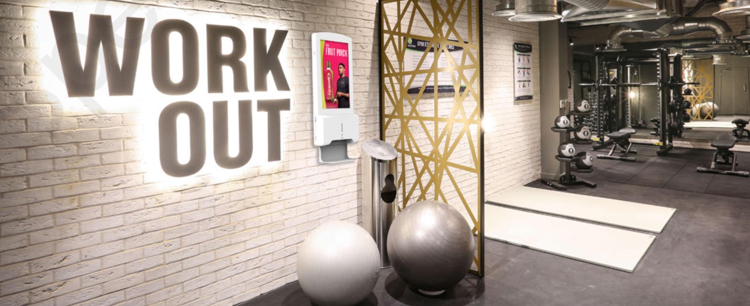 Digital hand sanitiser station mounted on a gym wall beneath a "WORK OUT" sign, positioned near the entrance to a fitness studio with equipment visible beyond.
