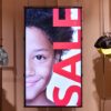 Vertical digital signage screen suspended in a children's clothing store, showing a bold "SALE" graphic overlaid on a portrait image alongside hanging garments.