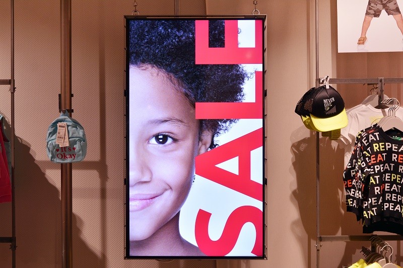 Vertical digital signage screen suspended in a children's clothing store, displaying a bold "SALE" graphic to drive in-store purchasing decisions.
