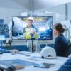 Manufacturing team gathered around a meeting table, viewing a wall-mounted display showing a colleague on a factory floor in full protective safety gear.