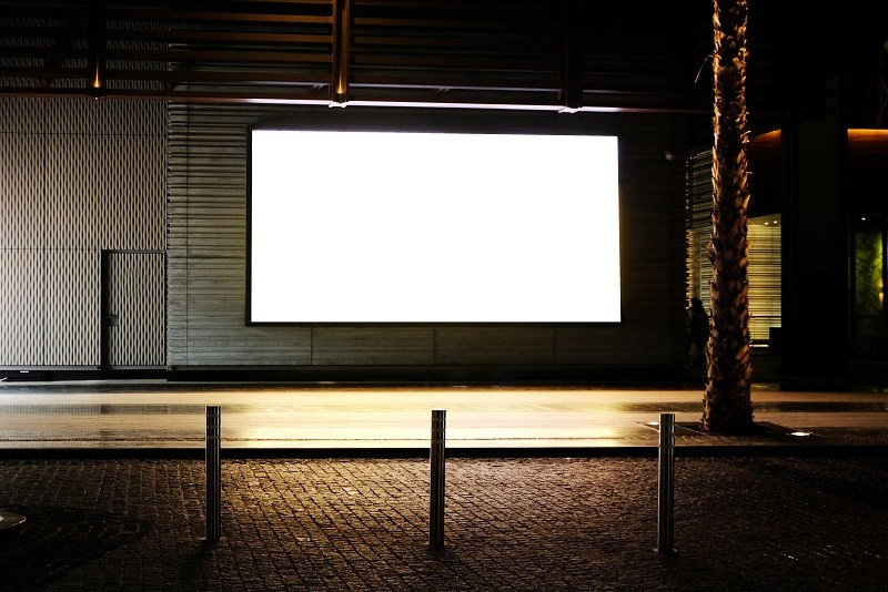 Blank illuminated digital signage screen mounted outdoors at night in a quiet, empty pedestrian area, showing no active content.