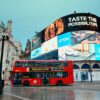 Large curved outdoor LED screen at Piccadilly Circus displaying multiple brand advertisements above a busy road with a red double-decker bus passing.