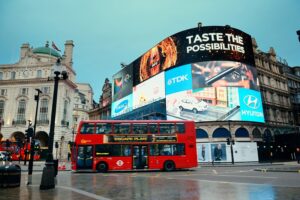 Large curved outdoor LED screen at Piccadilly Circus displaying multiple brand advertisements above a busy road with a red double-decker bus passing.