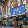Large suspended digital signage screen inside a railway station displaying an NHS public health message reminding travellers to protect each other.