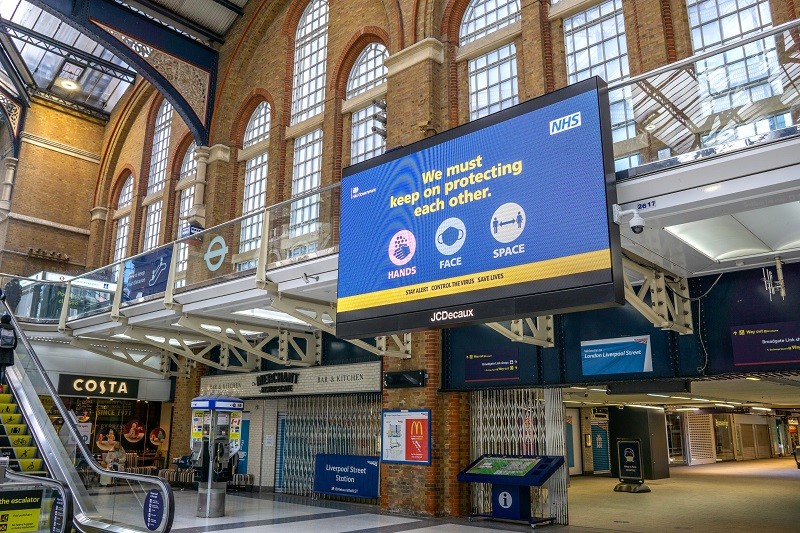 Large suspended digital signage screen inside a railway station displaying an NHS public health message reminding travellers to protect each other.