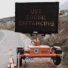 Portable trailer-mounted LED road sign displaying "USE SOCIAL DISTANCING" beside a rural roadside, with traffic cones marking the area.