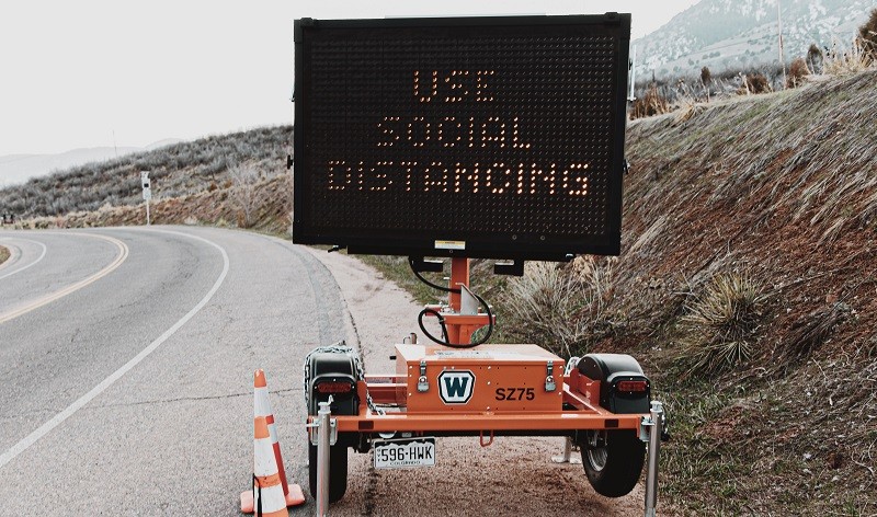 Portable trailer-mounted LED road sign displaying "USE SOCIAL DISTANCING" beside a rural roadside, with traffic cones marking the area.