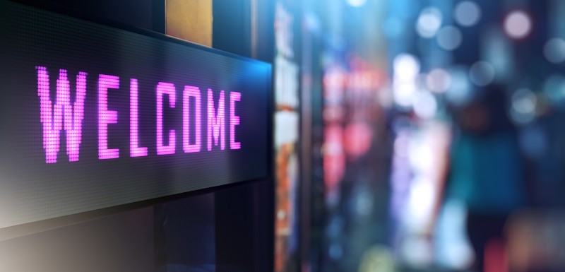Illuminated LED sign displaying "WELCOME" in pink text against a blurred, softly lit night-time background.