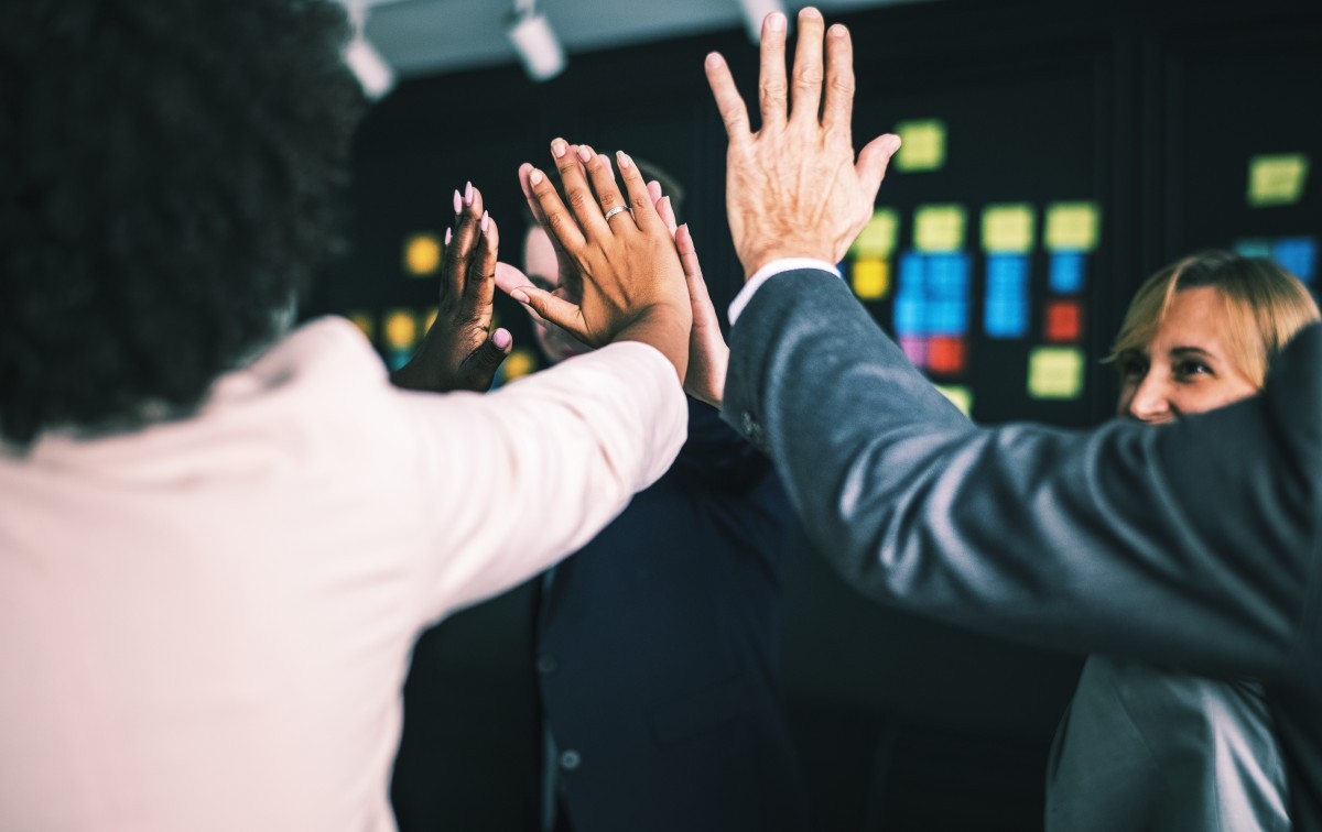 Colleagues raising hands together in a group high-five in front of a wall covered in colourful sticky notes, reflecting team collaboration and shared communication.