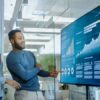 Colleague presenting data analysis on a wall-mounted digital display screen to two team members in a modern glass-walled office.