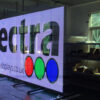Transparent LED screen displaying the Spectra logo in a workshop setting, with shelving and equipment visible through the open mesh structure behind it.
