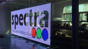 Transparent LED screen displaying the Spectra logo in a workshop setting, with shelving and equipment visible through the open mesh structure behind it.