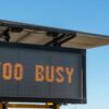 Portable outdoor LED sign displaying "TOO BUSY" against a clear blue sky, mounted on a trailer unit with a solar panel.