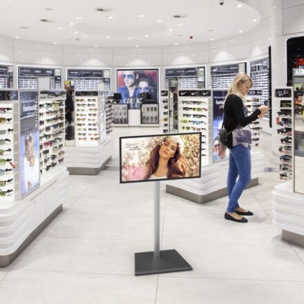 LC2246FP tilting floor stand holding a digital display showing fashion imagery, positioned in a sunglasses retail store as a customer browses nearby.