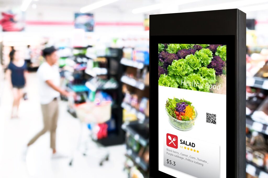 Freestanding digital signage totem displaying a salad promotion in a supermarket aisle, illustrating energy-efficient in-store advertising.