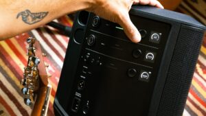 Musician adjusting the control panel of a Bose S1 Pro+ Wireless PA System, with a guitar positioned alongside for a live performance setup.