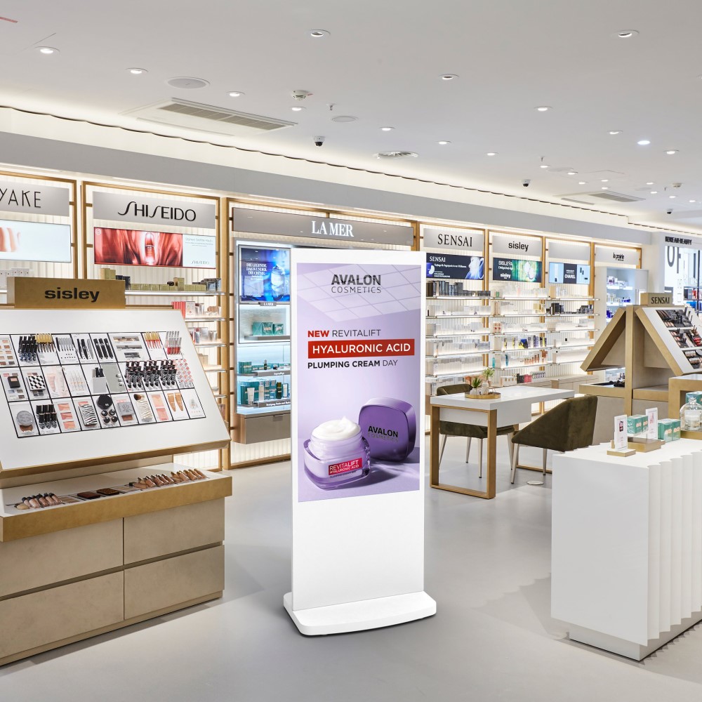 White 50-inch freestanding digital poster in a cosmetics department store, displaying a skincare product advert.