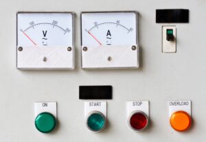 Industrial control panel showing voltage and current meters alongside start, stop, and overload push buttons.
