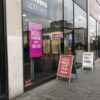 Opticians shopfront with a bright pink window display advertising hearing aids, alongside printed pavement signage for eye test offers.