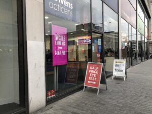 Opticians shopfront with a bright pink window display advertising hearing aids, alongside printed pavement signage for eye test offers.