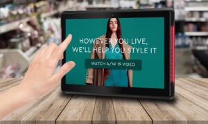 Interactive POS touchscreen in a clothing store displaying a seasonal fashion campaign with a watch video prompt.