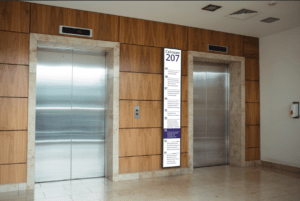 Ultra-wide stretched digital display showing floor directory information, mounted between two lift doors.