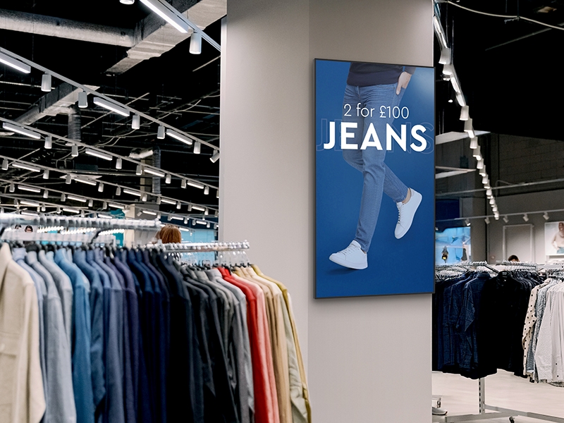 Vibrant professional digital advertising display screen mounted in portrait orientation on a pillar inside a high-street retail clothing shop, displaying an apparel promotional graphic.